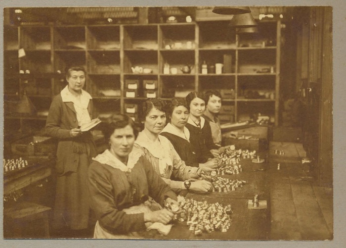 Five women sit at a workbench in front of piles of metal pieces. Another woman stands behind them