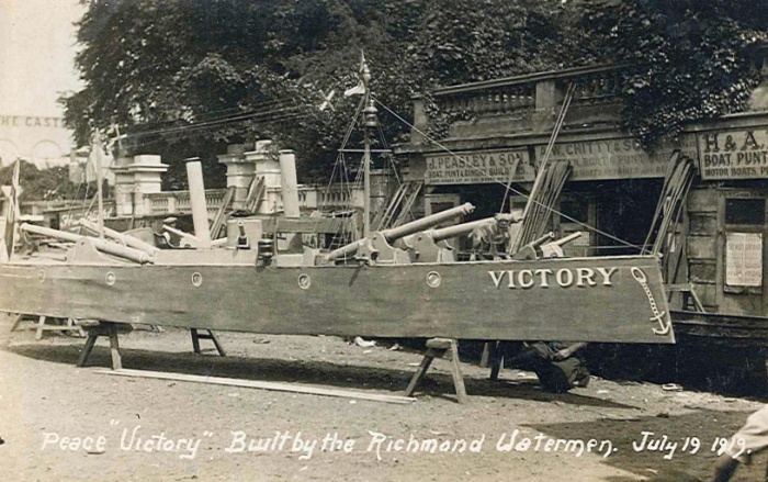 A rowing boat stands on the shore outside workshops. The handwritten caption reades: "Peace Victory. Built by the Richmond Watermen. July 19 1919