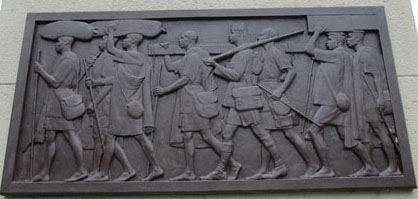 Carved bronze relief showing seven African men carrying sacks and boxes above their heads. The men wear a uniform with short trousers and all are barefoot.